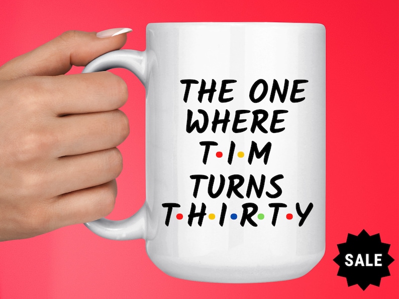 May include: White ceramic mug with black text that reads "The One Where Tim Turns Thirty" with colorful dots replacing the vowels. The mug has a black handle and is held by a hand.