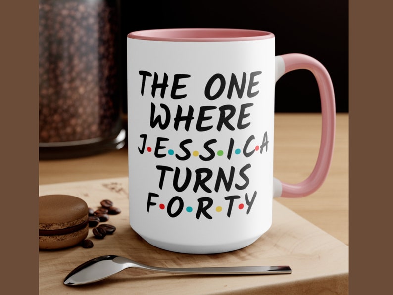 Op de afbeelding: Witte keramische mok met een roze rand en handvat. De mok heeft een zwarte afbeelding met de tekst "The One Where Jessica Turns Forty" met gekleurde stippen die de letters voorstellen.