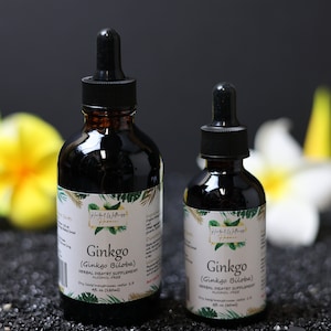Puede incluir: Dos botellas de vidrio marrón con goteros, etiquetadas "Ginkgo (Ginkgo Biloba) Suplemento dietético herbal sin alcohol 4 fl oz (120 ml)". Las botellas están sobre una superficie negra con flores amarillas en el fondo.