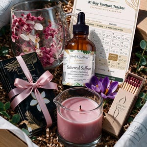 May include: A wellness gift set containing a glass mug filled with pink dried flowers, a brown glass dropper bottle labeled Satiereal Saffron, a 31-Day Tincture Tracker card, a black box with a pink ribbon, a pink candle, and a box of matches.