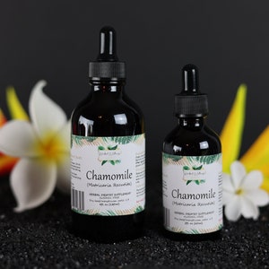 May include: Two bottles of chamomile herbal therapy supplement, one larger and one smaller, both with a dropper, on a black surface with yellow and white flowers in the background.
