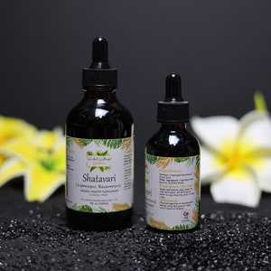 May include: Two bottles of Shatavari (Asparagus Racemosus) herbal extract, one larger and one smaller, with a white label and green foliage design. The bottles are on a black surface with yellow flowers in the background.