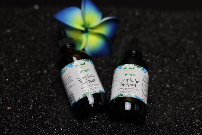 May include: Two brown glass bottles with a white label that reads "Lymphatic Support" on a black background. The bottles are surrounded by black sand and a blue and yellow flower.