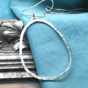 May include: A silver-toned, hammered-metal pendant necklace with an oval-shaped pendant. The pendant hangs from a delicate chain against a teal-colored fabric background. The necklace is displayed on a wooden surface.