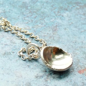 May include: A silver pendant necklace featuring a small, textured, open-faced shell design. The pendant hangs from a delicate silver chain. The background is a mottled blue and teal.