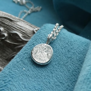 May include: A silver pendant necklace featuring an oval pendant with a textured surface. The pendant hangs from a fine silver chain. The necklace is presented on a blue velvet background, which highlights the silver's shine.