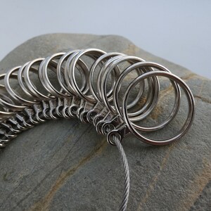 May include: A silver ring sizer with multiple rings of different sizes connected by a silver chain. The sizer is resting on a gray rock.