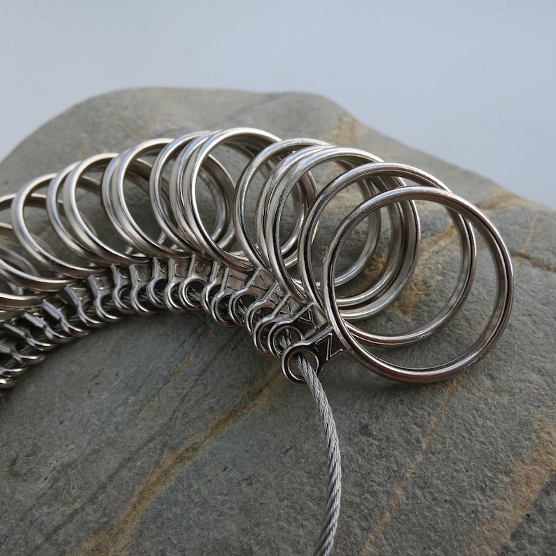 May include: A silver ring sizer with multiple rings of different sizes connected by a silver chain. The sizer is resting on a gray rock.