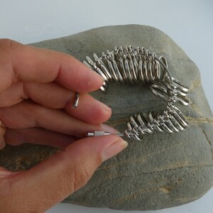 May include: A silver ring sizer with a cable attached. The sizer is made up of a series of connected rings of different sizes. The sizer is lying on a gray rock.