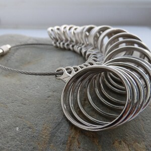 May include: A silver ring sizer with a chain and a clasp. The sizer has a series of rings of different sizes, each marked with a number.