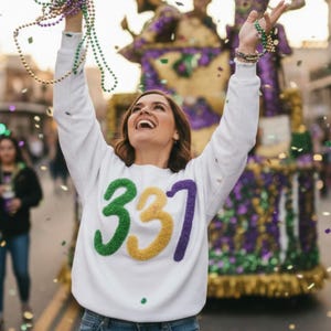 Op de afbeelding: Een vrouw in een witte trui met het nummer "337" in groen, geel en paars viert op een parade. Ze houdt kralen vast en confetti valt. Een praalwagen met figuren en versieringen staat op de achtergrond.