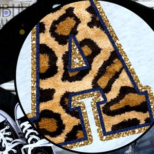 Puede incluir: Una camiseta negra de manga larga con una camiseta blanca con una gran letra "A" con diseño de estampado de leopardo. La letra está delineada en azul marino y purpurina dorada. Zapatillas de lona negras y vaqueros azules completan el conjunto.