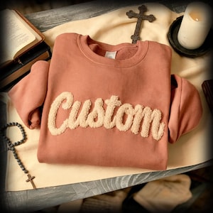 May include: A dusty rose sweatshirt with the word "Custom" in a fluffy, cream-colored font across the front. The sweatshirt is displayed on a cream-colored cloth, with a cross, rosary, and books in the background.