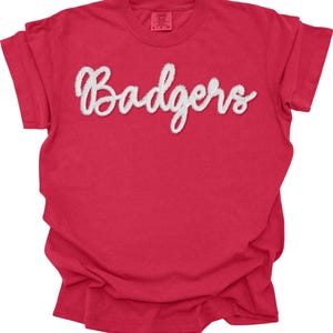 Könnte beinhalten: Rotes T-Shirt mit dem Wort "Badgers" in weißer, kursiver Schrift auf der Vorderseite. Das T-Shirt hat kurze Ärmel und einen Rundhalsausschnitt. Das Etikett ist oben am Rücken sichtbar. Der Stoff wirkt weich.