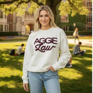 May include: Cream-colored sweatshirt with the text "Aggie Law" in maroon script. The sweatshirt is worn with blue jeans. The image is taken outdoors on a sunny day, with a university building in the background.