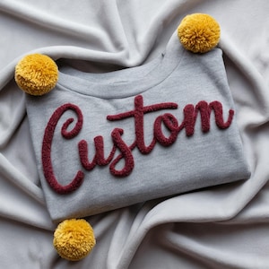 Peut inclure: Un sweat-shirt gris avec le mot "Custom" en bordeaux, avec une police texturée. Trois pompons jaunes sont placés autour du sweat-shirt sur un fond en tissu gris doux. Le sweat-shirt a un col rond.