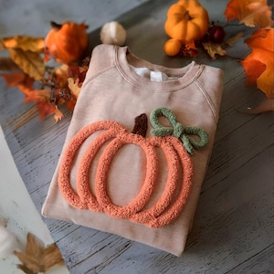 Peut inclure: Un sweat-shirt beige avec une grande citrouille orange texturée, ornée d'un nœud vert et d'une tige marron. L'image est agrémentée de feuilles d'automne et de mini-citrouilles, créant un thème saisonnier.