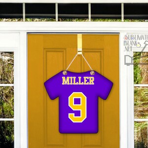 Puede incluir: Un colgador de puerta con forma de camiseta morada con el nombre "MILLER" y el n&uacute;mero "9" en amarillo. El colgador est&aacute; suspendido por una cadena plateada y cuelga de una puerta dorada.