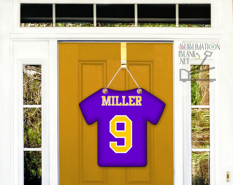 Puede incluir: Un colgador de puerta con forma de camiseta morada con el nombre "MILLER" y el n&uacute;mero "9" en amarillo. El colgador est&aacute; suspendido por una cadena plateada y cuelga de una puerta dorada.
