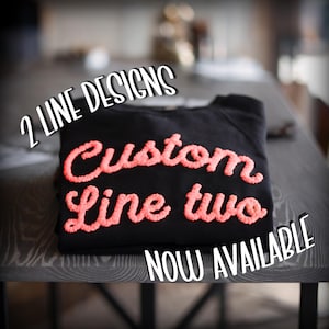 May include: A black sweatshirt featuring textured, coral-colored embroidered lettering that reads Custom Line two. The image includes the text 2 LINE DESIGNS and NOW AVAILABLE in white, bold, outlined font positioned above and below the garment.