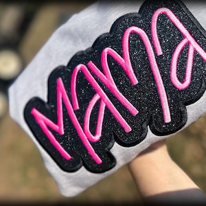 May include: A light gray sweatshirt with a large, glittery black patch that reads "MAMA" in bright pink script. The patch has a black border. The sweatshirt is held up against a blurred background.