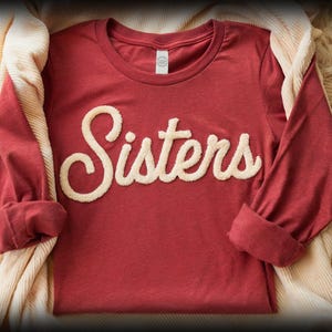 May include: A long-sleeved, red t-shirt with the word "Sisters" in a cream-colored, textured font across the front. The sleeves are cuffed, and the shirt is displayed on a cream-colored fabric and a fluffy, light-colored surface.
