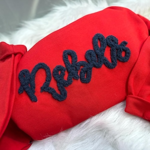 May include: A red sweatshirt with the word "Rebels" embroidered in navy blue script across the front. The sweatshirt is made of a soft material and is displayed on a white, fluffy surface.