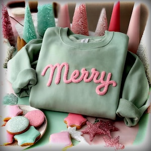 以下が含まれることがあります： ピンク色の「Merry」の文字が施された、セージグリーンのスウェットシャツ。さまざまな形と色の装飾されたクッキー、装飾的なクリスマスツリー、星のオーナメントが含まれています。全体的なテーマは、お祝いと休日のテーマです。
