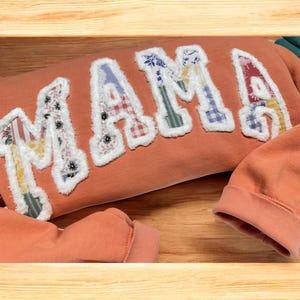 May include: An orange sweatshirt with the word "MAMA" appliquéd across the front. The letters are made of various patterned fabrics and outlined with white fluffy trim. The sleeves are visible at the bottom of the frame.