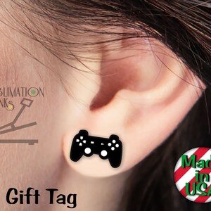 May include: Black stud earrings in the shape of a video game controller. The earrings are shown on an ear. The words "Gift Tag" are printed in black at the bottom of the image. The earrings are made in the USA.