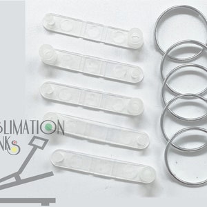 May include: A collection of clear plastic sublimation blanks and silver metal key rings. The plastic blanks are rectangular with rounded ends and small circular protrusions. The key rings are circular and made of shiny metal. The image includes the text "SUBLIMATION BLANKS .NET".