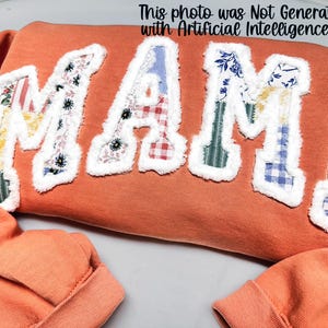 Op de afbeelding: Een oranje sweatshirt met het woord "MAMA" op de voorkant. De letters zijn gemaakt van verschillende stoffen met patronen, waaronder bloemen-, ruit- en streepmotieven, en zijn afgezet met witte, pluizige randen. De tekst "This photo was Not Generated with Artificial Intelligence!!" staat bovenaan.