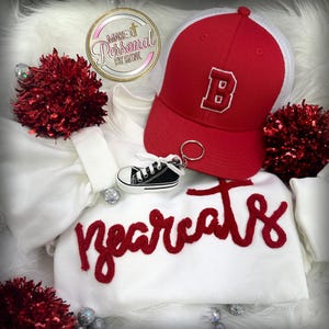 May include: A white sweatshirt with the word "Bearcats" in red script, a red baseball cap with a white mesh back and a red "B" patch, and a black and white sneaker keychain. The image also includes red pom-poms.
