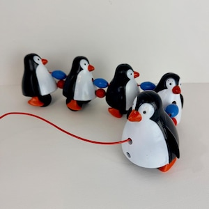 May include: A toy featuring five penguin figures in black, white, and orange, connected by blue and red beads. A red string extends from the lead penguin. The penguins have orange feet and beaks.