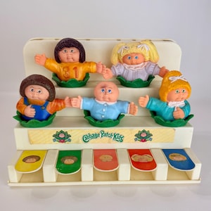Puede incluir: Un expositor de juguetes Cabbage Patch Kids con cinco pequeñas figuras de plástico que emergen de hojas de col verdes. El soporte tiene una base escalonada de color crema con cinco palancas coloridas en la parte inferior, cada una con una ilustración.