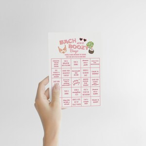 May include: A white card with pink text that reads "Bach and Boozy Bingo." The card features illustrations of a bikini top, a cocktail, and a tropical plant. The bingo card has various prompts for a bachelorette party.