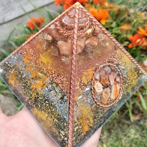 May include: A clear glass pyramid with copper wire wrapped around the edges. The pyramid is filled with various crystals and stones, including orange, brown, and gold pieces. The pyramid is sitting on a hand.