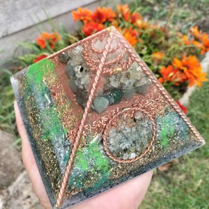 May include: A pyramid-shaped crystal grid with copper wire and various green and clear crystals. The grid is set on a dark grey base and surrounded by green and brown foliage.
