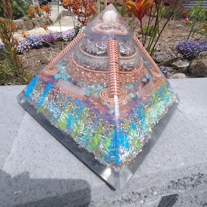 Puede incluir: Una pirámide clara y en capas con alambre de cobre, cristales y acentos coloridos. La pirámide presenta un diseño de varios niveles con piedras incrustadas y una parte superior puntiaguda. La base es transparente.