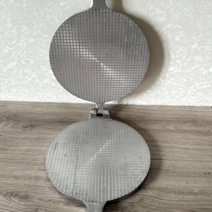 May include: A silver-colored metal waffle iron with two circular plates featuring a grid pattern. The device has a hinged design with long handles extending from the top and bottom, and black accents on the ends of the handles for gripping.