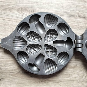 May include: A vintage, cast-iron cookie mold with various shell and nut-shaped cavities. The hinged mold, with a long handle, is made of gray metal and rests on a wooden surface. The mold is marked "USA".