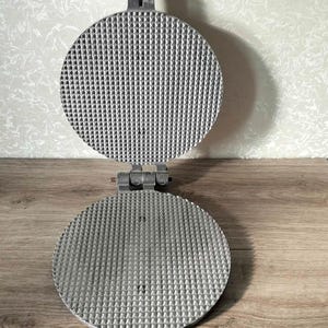 May include: A vintage, round waffle iron made of silver-colored metal. The iron is open, revealing two circular cooking surfaces with a textured, grid-like pattern. The handle extends upwards, and the iron is hinged in the middle. The background is a light wood surface.
