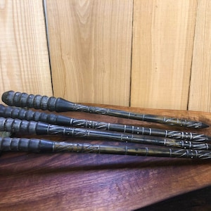 May include: Five black wooden wands with intricate carvings and silver accents. The wands are arranged in a row on a wooden surface.