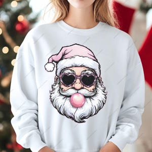May include: A white sweatshirt with a pink and white Santa Claus wearing heart-shaped sunglasses and blowing a pink bubble. The Santa Claus has a white beard and mustache.