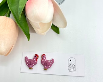 May include: A pair of pink and white polka dot chicken earrings. The earrings are handmade and feature a red comb and wattle. The earrings are on a white card with the text "Tyn + Bee Handmade".
