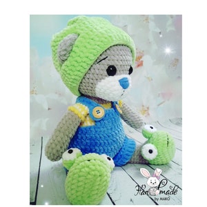 Puede incluir: Un oso de peluche hecho a mano con ganchillo, con un gorro verde, un peto azul y una camisa amarilla. El oso tiene pelaje gris, una nariz azul y está sentado con pies verdes con forma de rana. La imagen incluye el texto "Handmade by MAKO".