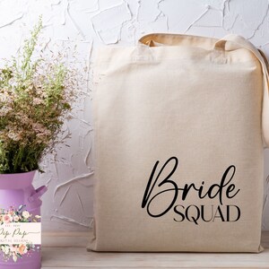 Op de afbeelding: Een beige canvas draagtas met de woorden "Bride Squad" in zwart script. De tas heeft lange handvatten. Daarnaast staat een paarse melkkanvaas met droogbloemen en een bloemenlabel.