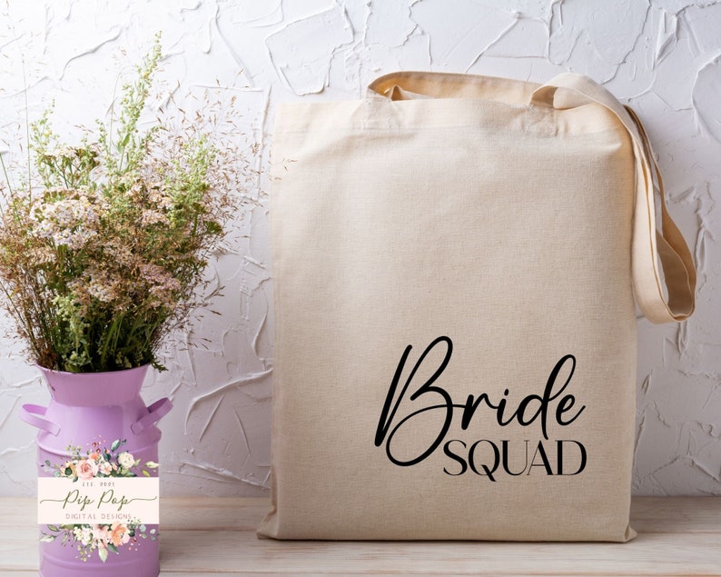 Op de afbeelding: Een beige canvas draagtas met de woorden "Bride Squad" in zwart script. De tas heeft lange handvatten. Daarnaast staat een paarse melkkanvaas met droogbloemen en een bloemenlabel.