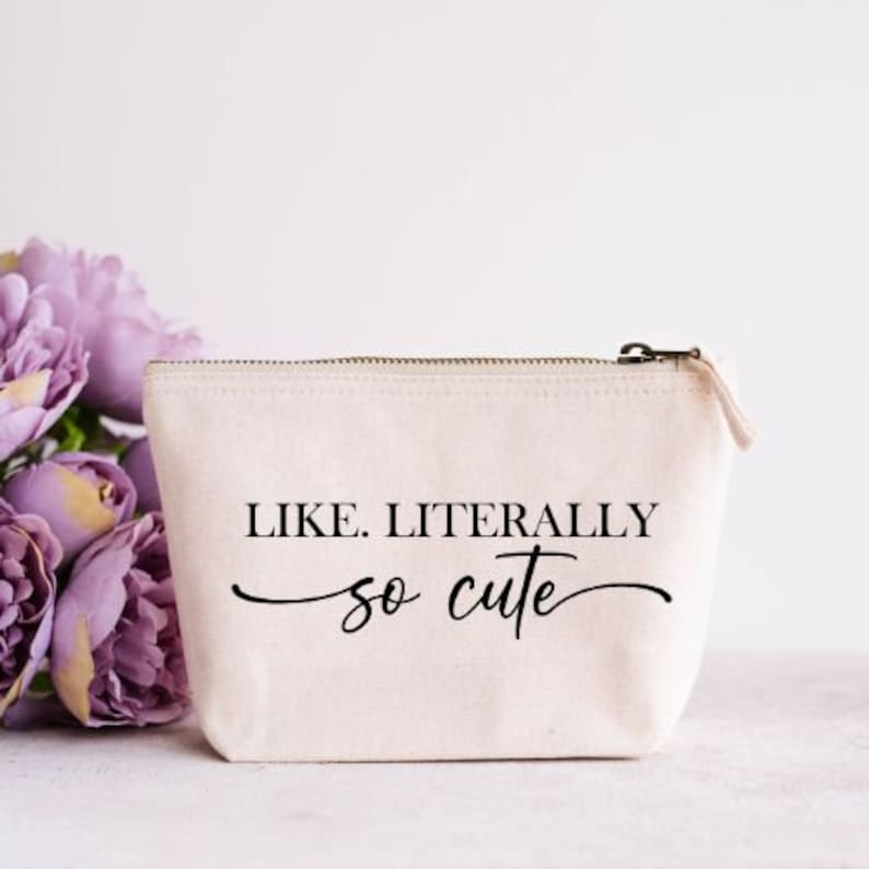 May include: A beige canvas cosmetic bag with a zipper closure. The bag features the text "LIKE. LITERALLY so cute" in black script. A bouquet of purple flowers is in the background.