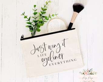 May include: A white canvas makeup bag with a black zipper and black text that reads "Just wing it life eyeliner everything".